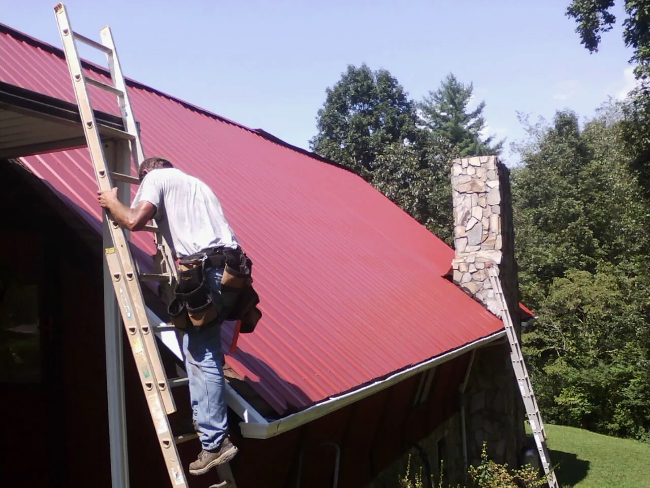 Roofer on a ladder beside a red metal roof and stone chimney — Soffit & Fascia Repair by Roofing Brothers in Smallwood